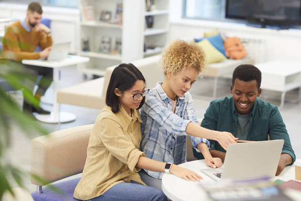 Visão de alto ângulo em grupo multi-étnico de estudantes alegres usando laptop juntos enquanto trabalham no projeto na biblioteca, espaço de cópia
 - Foto, Imagem