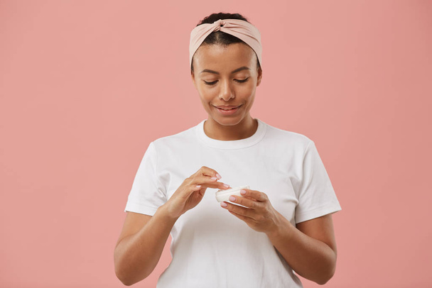 Porträt einer Mixed-Race-jungen Frau, die Gesichtscreme aufträgt und lächelt, während sie vor rosa Hintergrund während der morgendlichen Beauty-Routine posiert - Foto, Bild