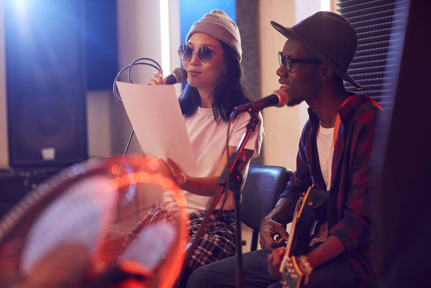 Çağdaş Afro-Amerikalı adamın gitar çalıp mikrofona şarkı söylediği resim. Prova veya konser sırasında, loş ışıklandırılmış stüdyo ortamında, Asyalı genç bir kadınla. - Fotoğraf, Görsel