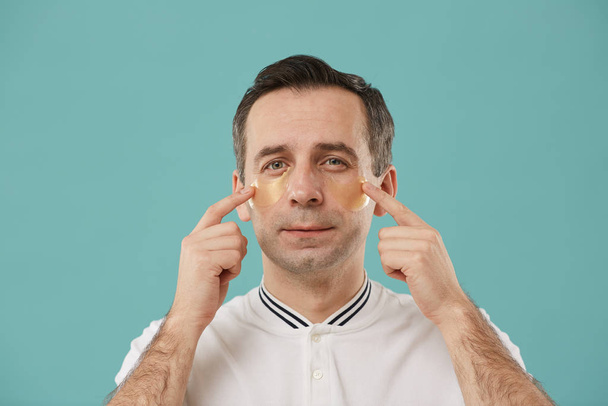 Head and shoulders portrait of middle aged man applying eye mask and looking at camera while enjoying beauty routine standing against blue background - Photo, Image