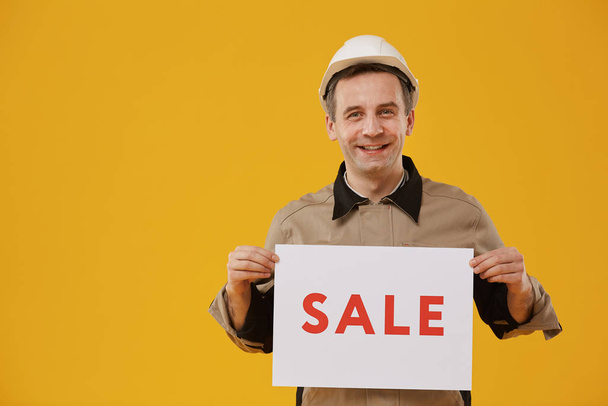 Waist up portrait of cheerful mature worker holding SALE sign standing against yellow background and smiling at camera, copy space - Фото, зображення