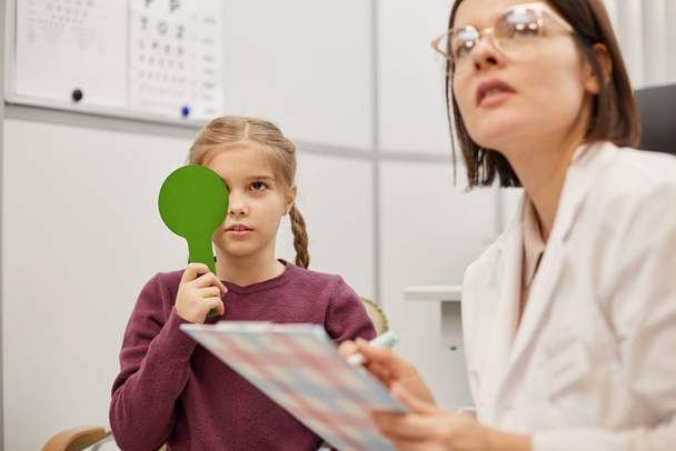 Ritratto di carina bambina che legge il grafico durante il test visivo nella clinica di oftalmologia pediatrica, spazio per la copia
 - Foto, immagini
