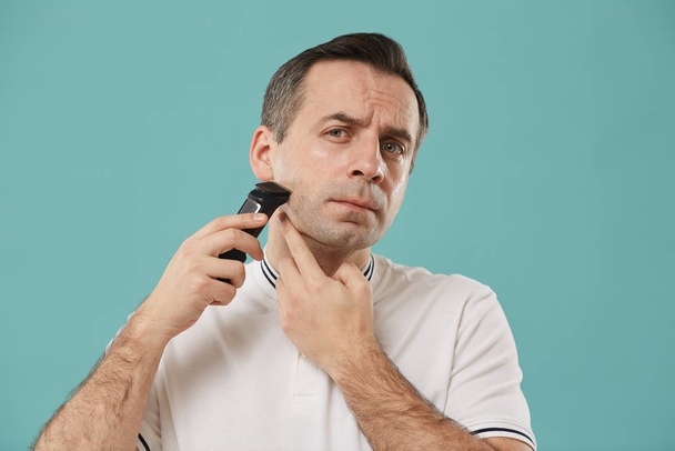 Head and shoulders portrait of handsome middle aged man shaving and looking at camera while enjoying morning routine standing against blue background - Photo, Image