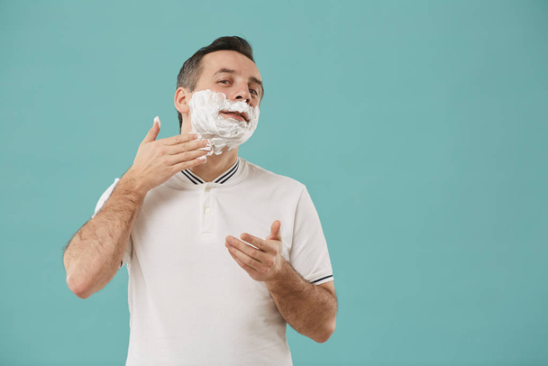 Waist up portrait of modern adult man shaving and looking at camera while enjoying morning routine standing against blue background, copy space - Foto, afbeelding