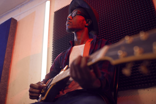 Low-Winkel-Porträt eines zeitgenössischen afroamerikanischen Mannes, der Gitarre spielt, auf einem Hochstuhl sitzend, während er im schummrig beleuchteten Studio Musik schreibt, Kopierraum - Foto, Bild