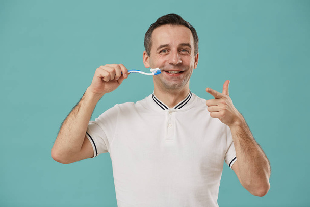 Cintura para cima retrato de homem adulto sorridente segurando escova de dentes e apontando para a câmera enquanto desfruta de rotina matinal em pé contra fundo azul
 - Foto, Imagem