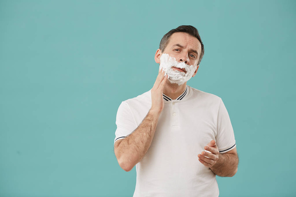 Waist up portrait of modern middle aged man shaving and looking at camera while enjoying morning routine standing against blue background, copy space - Fotó, kép