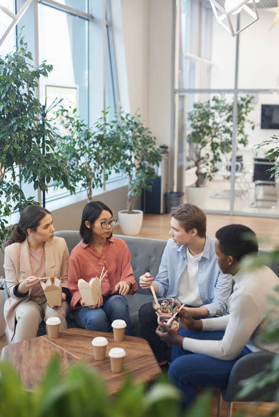 Высокоуровневый взгляд на многонациональную группу молодых людей, которые едят еду на вынос и наслаждаются беседой во время обеденного перерыва в офисе, копировальное пространство
 - Фото, изображение