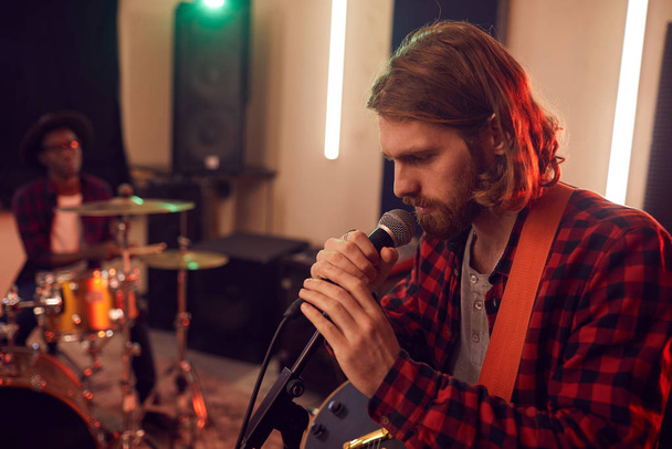Prova sırasında mikrofona şarkı söyleyen yakışıklı sakallı bir adamın yan görüntüsü ya da loş bir stüdyoda müzik grubuyla konser vermesi. - Fotoğraf, Görsel