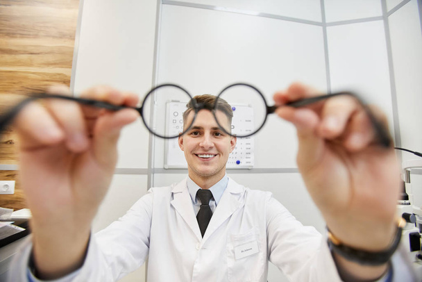 Фотография улыбающегося молодого оптометриста, надевающего очки на неузнаваемого пациента во время теста зрения в современной клинике, копировальное пространство
 - Фото, изображение