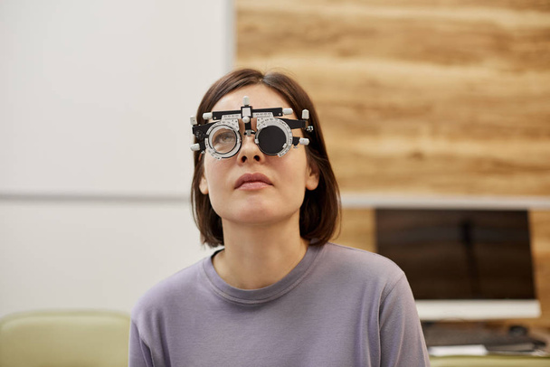 Head and shoulders portrait of young woman wearing trial frame during vision test in ophthalmology clinic, copy space - Foto, afbeelding