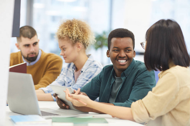 Grupo multi-étnico de estudantes que estudam na biblioteca universitária, com foco no homem afro-americano sorrindo para o parceiro
 - Foto, Imagem