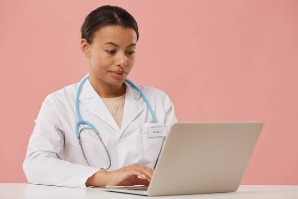 美しい女性医師の肖像ノートパソコンを使用しながら、薄いピンクの背景に座って、コピースペース - 写真・画像
