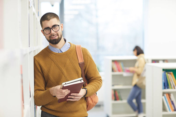 Waist up portrait of male student wearing glasses and looking at camera while posing by shelves in college library, copy space - Фото, зображення