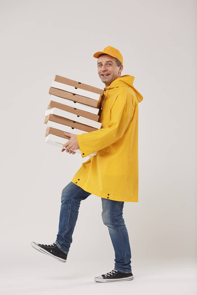 Side view full length portrait of adult delivery man holding stack of pizza boxes and smiling at camera walking against white background - Photo, Image