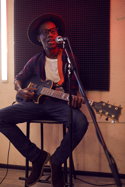 Full length portrait of contemporary African-American man playing guitar and singing to microphone while sitting on high chair during rehearsal or concert in dimly lit studio.. - Zdjęcie, obraz