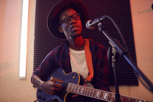 Портрет современного афроамериканца, играющего на гитаре и поющего под микрофон во время репетиции или концерта в тускло освещенной студии
 - Фото, изображение