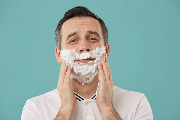Head and shoulders portrait of smiling adult man shaving and looking at camera while enjoying morning routine standing against blue background, copy space - Foto, Imagen