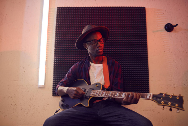 Weitwinkelporträt eines zeitgenössischen Afroamerikaners, der auf einem Hochstuhl sitzend Gitarre spielt, während er in einem schwach beleuchteten Studio Musik schreibt, Kopierraum - Foto, Bild