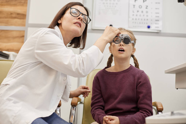 Modern oftalmoloji kliniğinde çocuk görme testi sırasında deneme çerçevesi takan sevimli genç kız portresi - Fotoğraf, Görsel