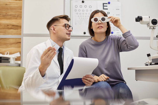 Portrait of young optometrist talking to young woman during vision test in modern ophthalmology clinic, copy space - Foto, Imagem