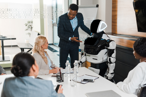 handsome african american businessman pointing with hand at robot near multicultural colleagues in  conference hall - Photo, Image