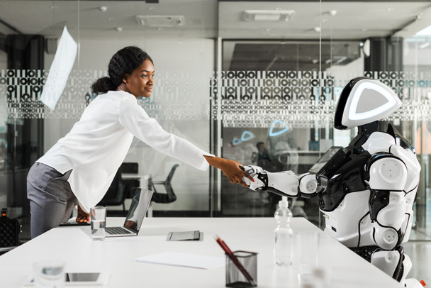 χαμογελαστή αφροαμερικανίδα επιχειρηματίας που σφίγγει το χέρι με ρομπότ στο γραφείο - Φωτογραφία, εικόνα