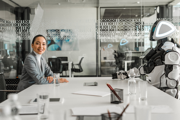 selektywne skupienie się uśmiechniętej azjatyckiej bizneswoman patrząc w kamerę siedząc przy biurku w pobliżu robota - Zdjęcie, obraz