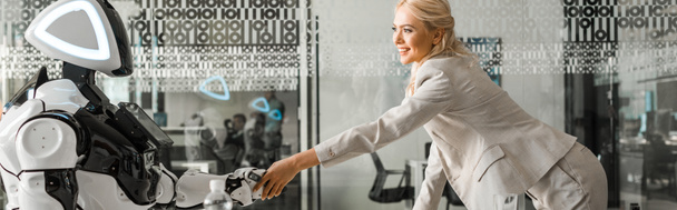 Панорамный снимок улыбающейся деловой женщины, пожимающей руку роботу, сидящему за столом в конференц-зале
 - Фото, изображение