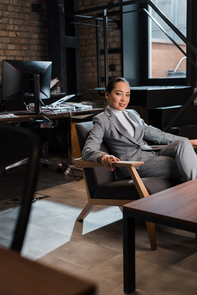 beautiful asian businesswoman sitting in armchair and smiling at camera - Fotografie, Obrázek
