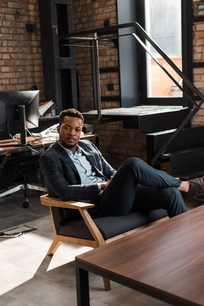 pohledný, sebevědomý africký americký podnikatel sedící v křesle a dívající se do kamery - Fotografie, Obrázek