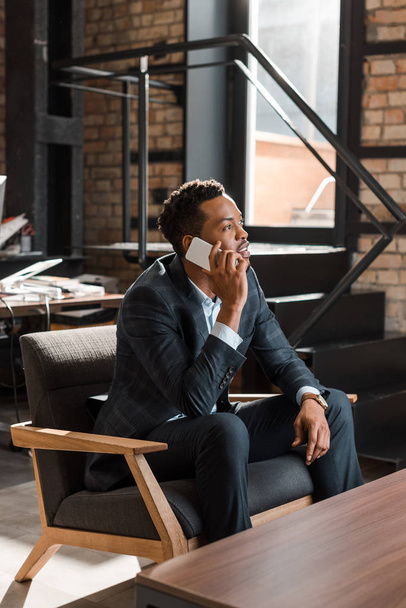 töprengő afro-amerikai üzletember ül karosszékben, beszél okostelefonon, és félrenéz  - Fotó, kép