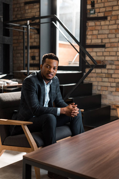 hombre de negocios afroamericano positivo sentado en sillón y sonriendo a la cámara
 - Foto, imagen