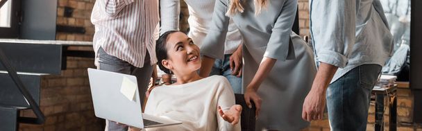 cropped view of multicultural businesspeople looking at cheerful asian colleague sitting with laptop - Foto, Bild