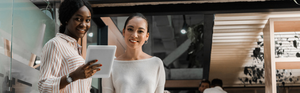 plano panorámico de la sonriente mujer de negocios afroamericana sosteniendo tableta digital mientras está cerca de colega
 - Foto, Imagen