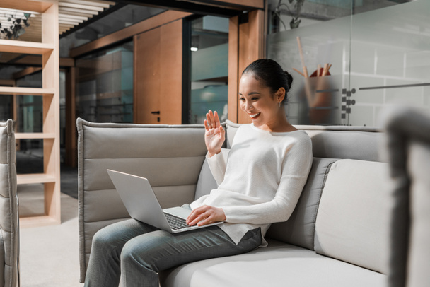 heureux asiatique femme d'affaires agitant la main tout en étant assis sur canapé et avoir chat vidéo sur ordinateur portable
 - Photo, image