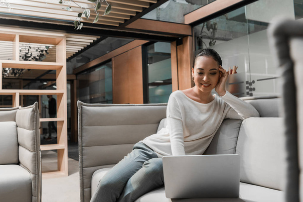 вибірковий фокус усміхненої азіатської бізнес-леді сидить на дивані і використовує ноутбук
 - Фото, зображення