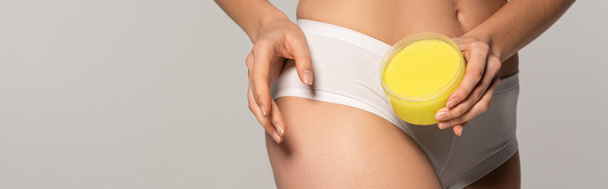 πανοραμική λήψη της γυναίκας εφαρμογή κίτρινο αλάτι scrub από πλαστικό δοχείο, απομονώνονται σε γκρι  - Φωτογραφία, εικόνα