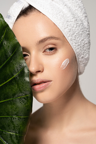 beautiful girl with cosmetic cream on face and towel on head posing with green leaf, isolated on grey  - Фото, изображение
