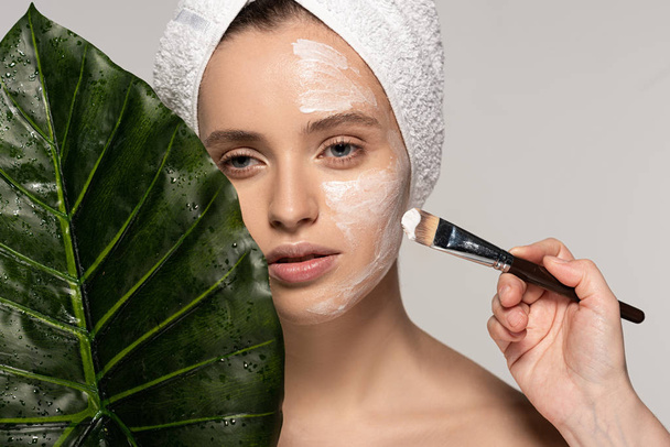aantrekkelijk meisje aanbrengen masker op gezicht met cosmetische borstel, geïsoleerd op grijs met blad - Foto, afbeelding