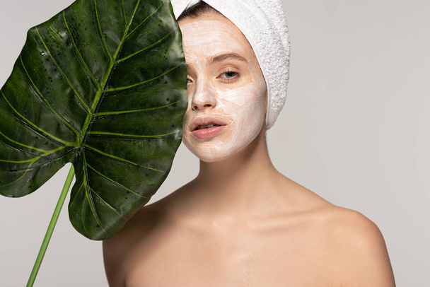 beautiful woman with cosmetic mask on face and towel on head posing with green leaf, isolated on grey  - Photo, Image