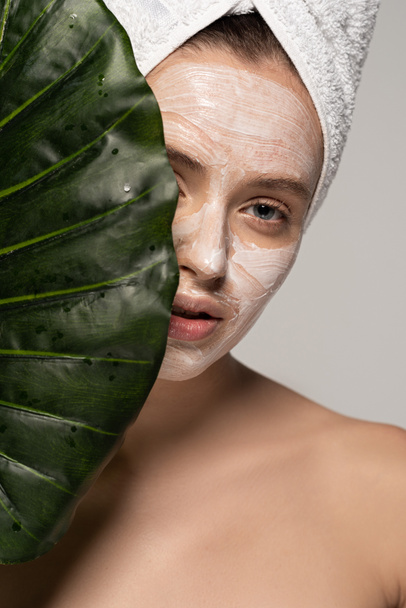 vonzó fiatal nő kozmetikai maszk az arcon és törölköző a fejen pózol zöld levél, elszigetelt szürke  - Fotó, kép