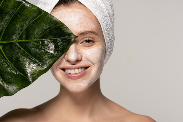 glückliche junge Frau mit Kosmetikmaske im Gesicht und Handtuch auf dem Kopf posiert mit grünem Blatt, isoliert auf grau  - Foto, Bild