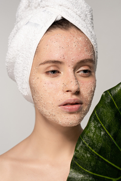 attractive girl with towel on head and coconut scrub on face posing with leaf, isolated on grey - Photo, Image