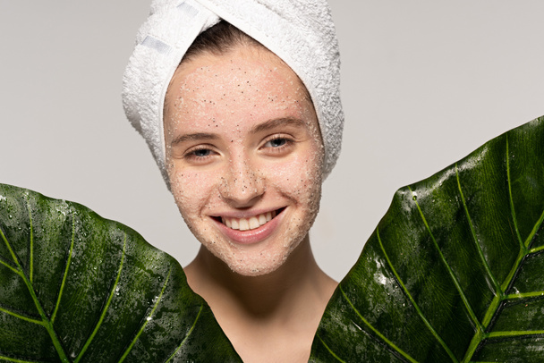uśmiechnięta dziewczyna z ręcznikiem na głowie i kokosowym peelingiem na twarzy pozująca z liśćmi, odizolowana na szaro - Zdjęcie, obraz