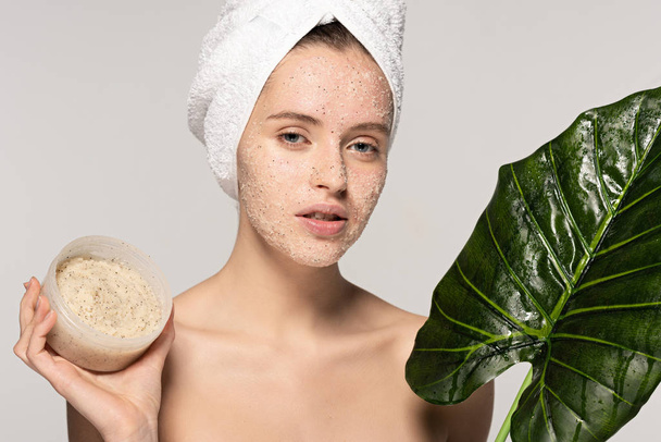 attractive girl with towel on head holding green leaf and plastic container with coconut scrub, isolated on grey - Photo, Image