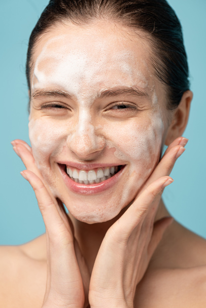 beautiful smiling young woman applying cleansing foam on face, isolated on blue - Foto, Imagen