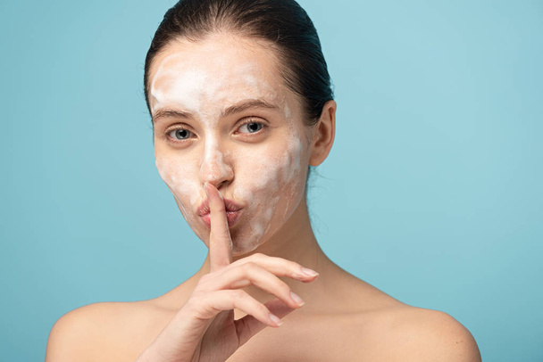 beautiful girl with cleansing foam on face showing silence symbol, isolated on blue - Photo, Image