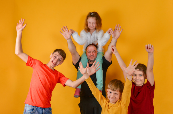Ένας πατέρας και τέσσερα χαρούμενα παιδιά σήκωσαν τα χέρια ψηλά, μια κόρη κάθεται στους ώμους του πατέρα μου. Αστεία και αστεία όμορφα παιδιά με πολύχρωμα ρούχα. - Φωτογραφία, εικόνα