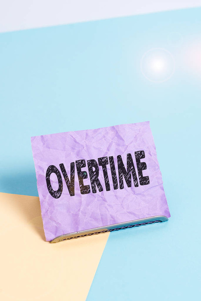 Handwriting text Overtime. Concept meaning Time or hours worked in addition to regular working hours Paper placed tilted above buffer wire on soft pastel multi colours backdrop. - Photo, Image